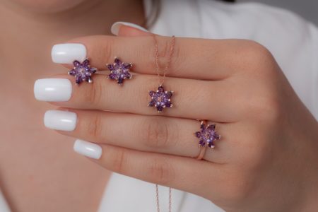 The young woman in a white dress and with a white nail polish hand shows the ring, necklace and earrings in the shape of a purple lotus flower in rose color