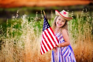 Happy adorable little girl smiling and waving American flag outside, her dress with strip and stars, cowboy hat. Smiling child celebrating 4th july - Independence Day